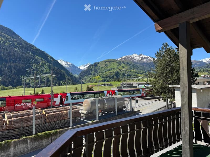 Balcony overlooking a scenic mountain landscape with a train station and passing train in the distance.