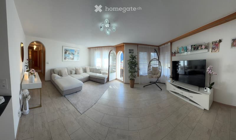 Modern living room with a white sectional sofa, coffee table, flat-screen TV on a white stand, wicker hanging chair, and decorative vases. Large windows with sheer curtains allow natural light to fill the space.