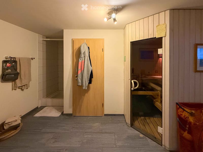 Bathroom with tiled walls, a shower stall, a wooden door with a hanging bathrobe, a towel rack with towels, and an open sauna area with wooden benches.