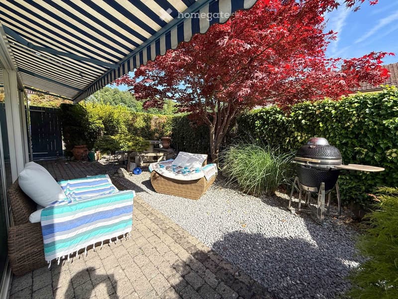 Outdoor patio with a striped awning, wicker sofa and chair, a round table, and a grill, surrounded by greenery and a red-leaved tree.