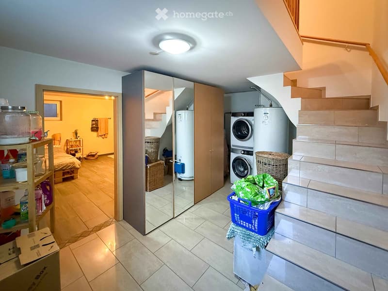 Hallway with tiled floor, staircase leading upwards, washing machine, water heater, and a partially open closet with a mirror. Laundry items and cleaning supplies are visible.