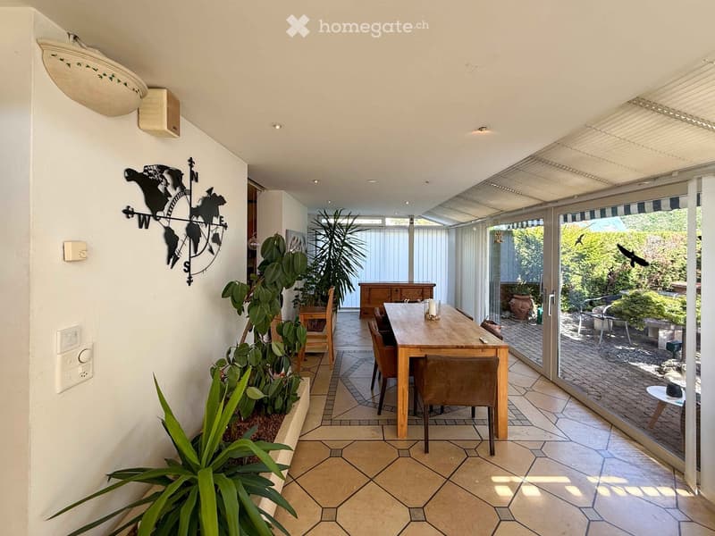 Bright, tiled dining area with a wooden dining table, chairs, and large potted plants. Large sliding glass doors open to a patio with outdoor furniture.