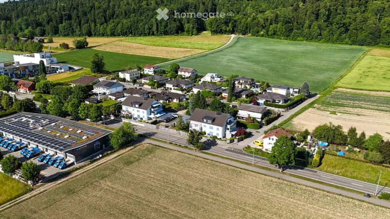 Aerial view of a residential area with multiple houses, a parking lot, and a swimming pool surrounded by greenery and farmland.