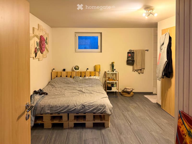 Cozy bedroom with a wooden pallet bed, nightstand with lamp, wall art, and a small shelf with decorative items. Wooden floor and neutral-colored walls.