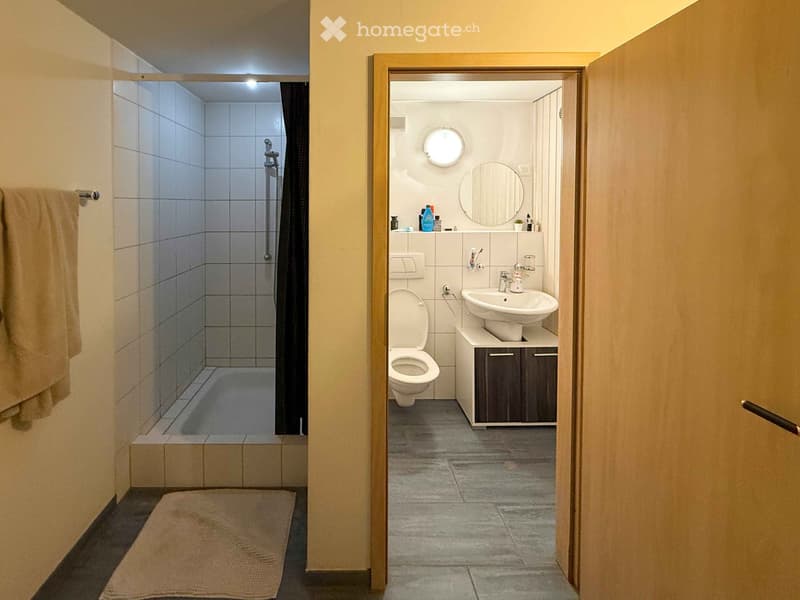Modern bathroom with a bathtub, toilet, sink, and mirror. Features tiled walls, a wooden cabinet, and a towel on the floor.