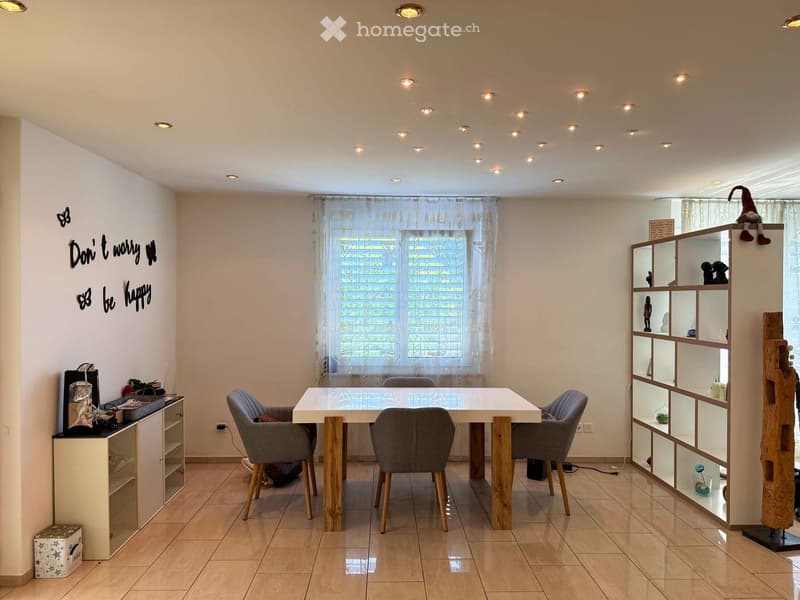 Bright dining area with a white table, four grey chairs, a wooden shelf unit, and a small storage cabinet. The room features a tiled floor, large windows with sheer curtains, and decorative wall art.