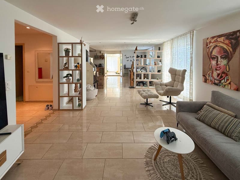 Modern living room with tiled flooring, large windows, a grey sofa, coffee table, armchair, and decorative shelves.