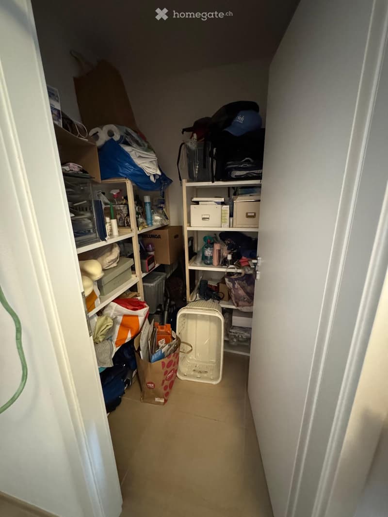 A narrow hallway with shelves on both sides filled with various items including boxes, bags, and storage containers. The floor is tiled and the walls are painted white.