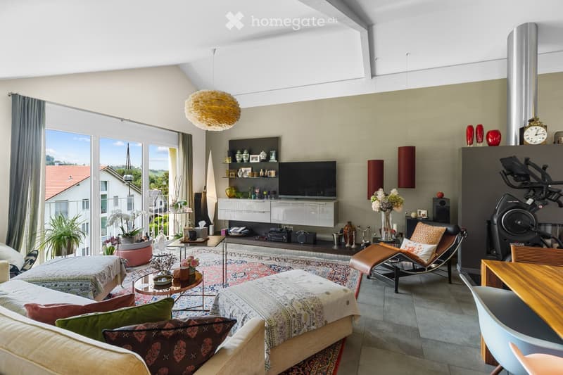 Modern living room with large windows offering a view, featuring a sofa, coffee table, ottoman, chair, exercise bike, shelves with decorative items, and a television.