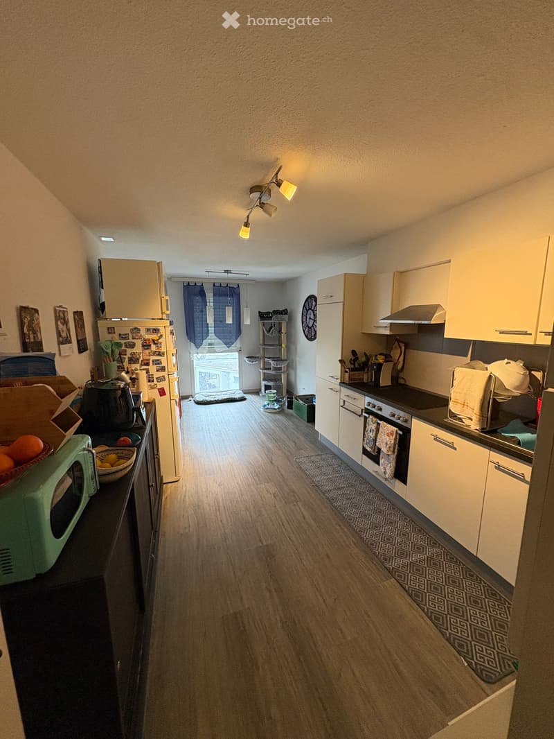 Modern kitchen with white cabinets, wooden flooring, and various appliances including a refrigerator, oven, microwave, and kitchen island. There is a window with blue curtains and a small pet area visible in the background.