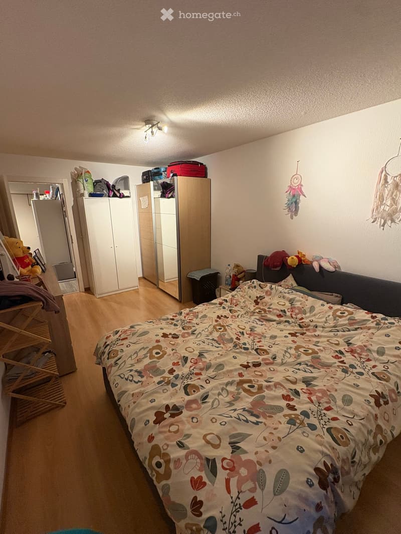 A cozy bedroom with a large bed covered in a floral duvet, a white wardrobe, a mirror, a wooden chair with a teddy bear, and various toys scattered around.
