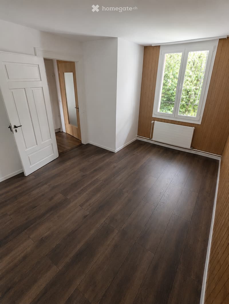 Unfurnished Room. Empty room with dark wooden flooring, white walls, and a white door leading to another room. There is a radiator under a window with wooden paneling on one wall.