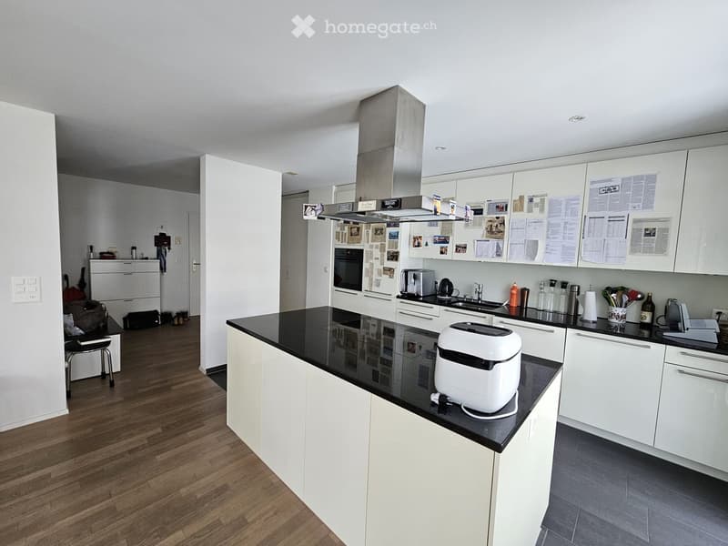 Modern kitchen with white cabinets, black countertops, and various appliances including a refrigerator, oven, sink, coffee maker, and toaster. Wooden flooring and a small stool near the refrigerator.
