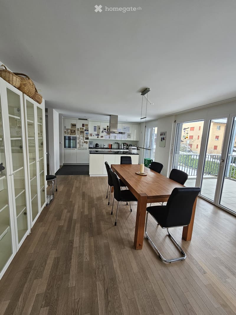 Open-plan dining and kitchen area with wooden flooring, a long wooden dining table with six chairs, a modern kitchen with white cabinets, an oven, refrigerator, and a kitchen island.