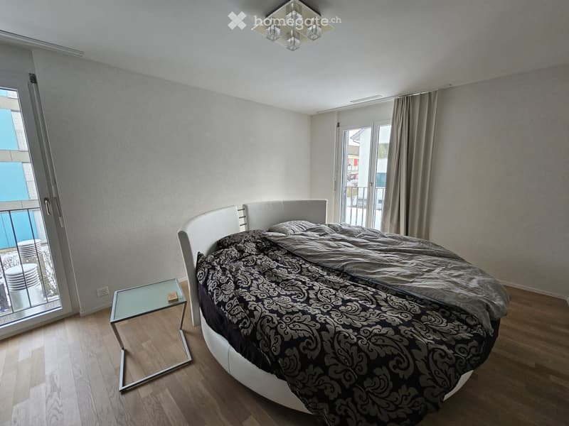 Modern bedroom with a large bed, patterned bedding, a glass nightstand, and a wooden floor. Large windows and sliding doors lead to a balcony.