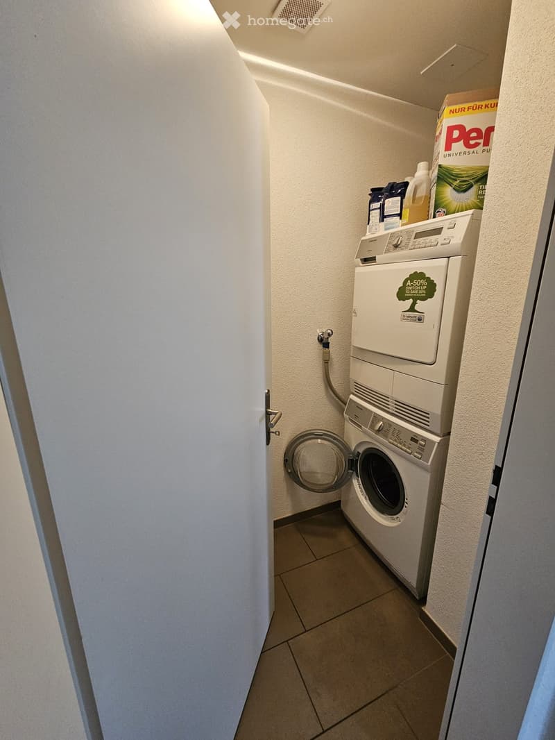 Compact bathroom with a washing machine and dishwasher stacked vertically, tiled floor, and cleaning supplies on top.