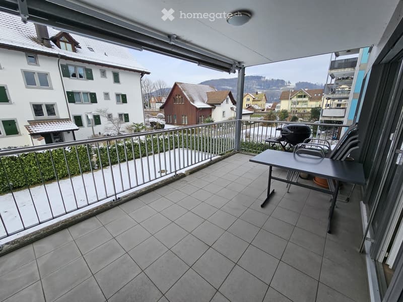 Balcony with tiled floor, metal railing, table, chairs, and grill, overlooking a residential area with houses and greenery.