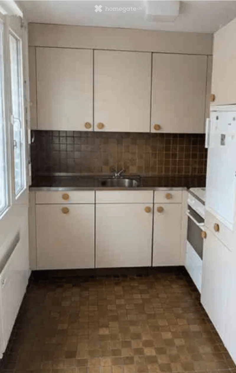 A clean, unfurnished kitchen with light-colored cabinets, a sink, and a refrigerator. The floor is tiled, and the backsplash features a dark, textured pattern.