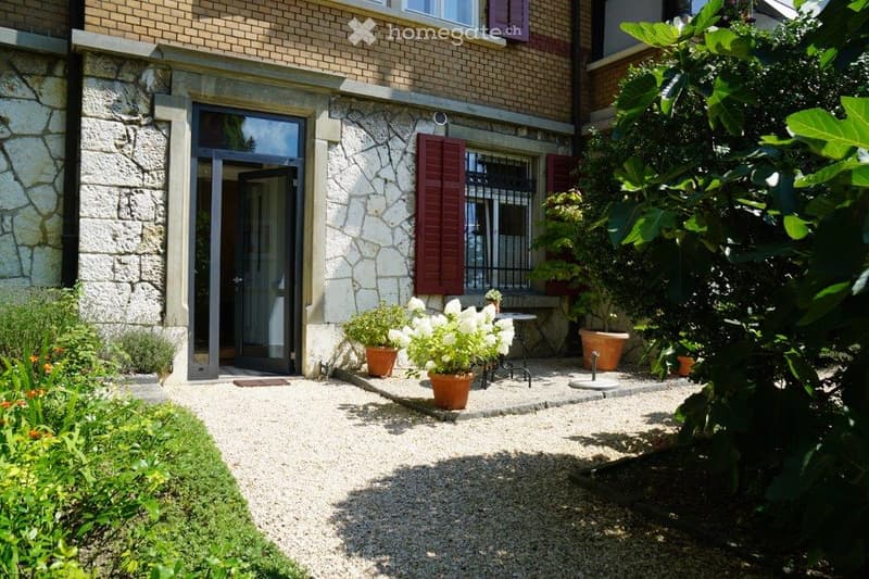 Garden area with gravel pathway, potted plants, small table, and bench near a stone house entrance.