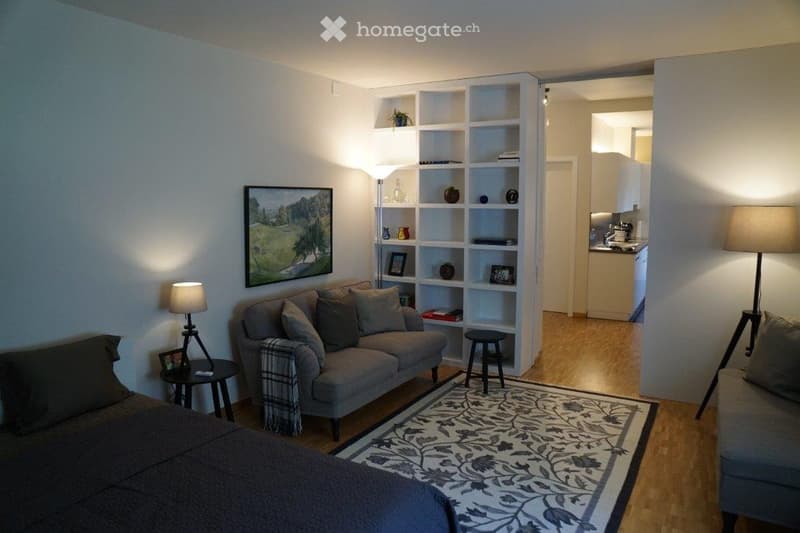 A cozy, well-furnished living space with a sofa, bed, small table, lamps, shelves, and a stool. The room features a patterned rug and a view into an adjacent kitchen area.