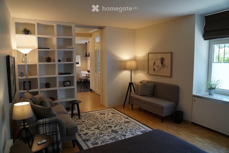 A cozy living room with a grey sofa, coffee table, floor lamp, bookshelf, and a small dining area visible in the background.