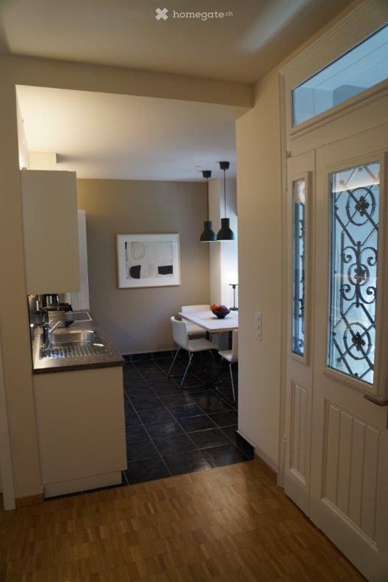 A modern, open-plan kitchen and dining area with light-colored cabinets, a stainless steel sink, a small dining table with chairs, and a framed artwork on the wall. The space features wooden flooring and pendant lights.