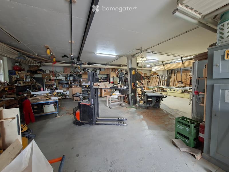 A spacious workshop with concrete floors, various tools and equipment, including a workbench, table saw, shelves, and a forklift.