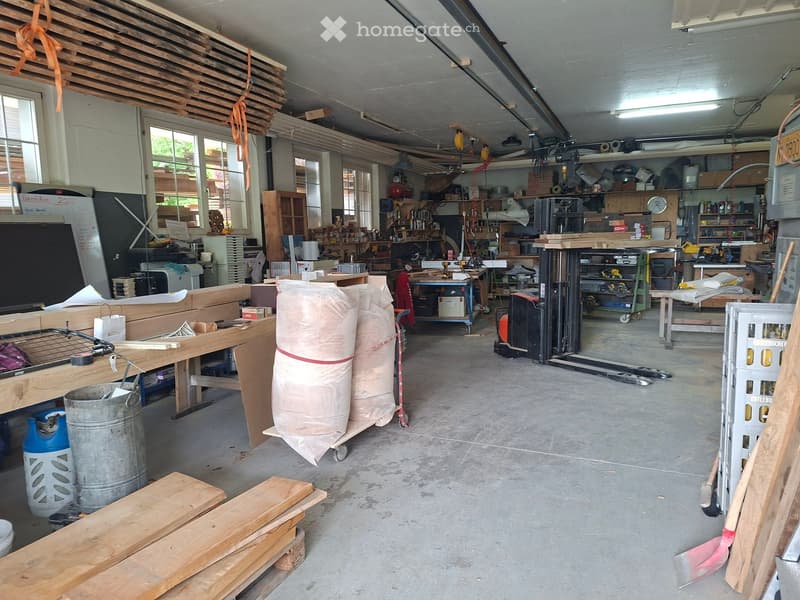 A spacious workshop with exposed wooden beams, large windows, and various tools and materials. Visible items include workbenches, shelves, wooden planks, barrels, and storage bins.