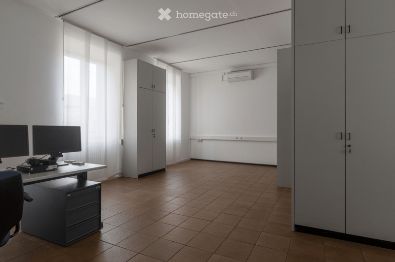 Modern office space with a tiled floor, large windows with white curtains, a desk with two computer monitors, a chair, a cabinet, and an air conditioner mounted on the wall.