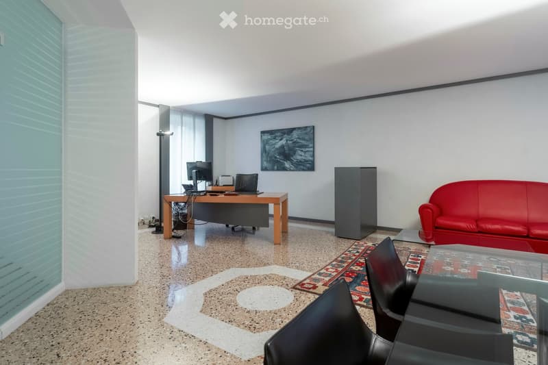 Modern office space with a red sofa, black leather chairs, a wooden desk with a laptop and screen, a glass coffee table, and a large painting on the wall.