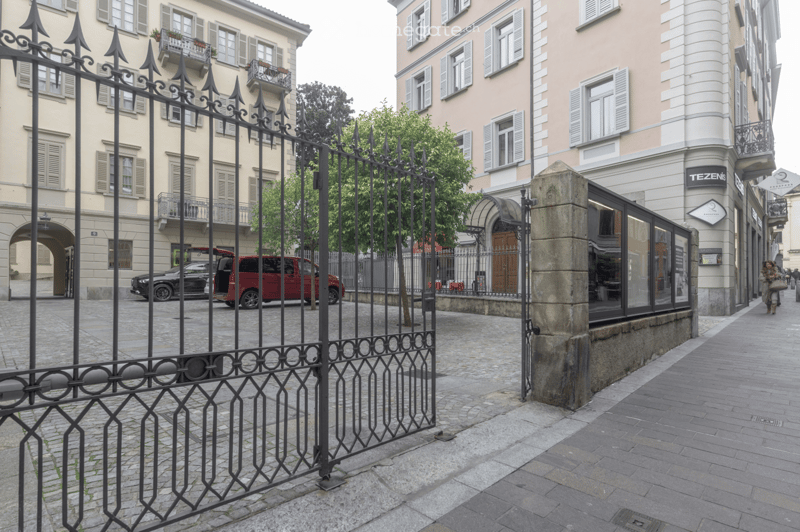 A narrow cobblestone street lined with multi-story buildings, featuring ornate wrought-iron fences, balconies, and a small tree. A red van is parked on the left, and a modern storefront with large glass windows is visible on the right.