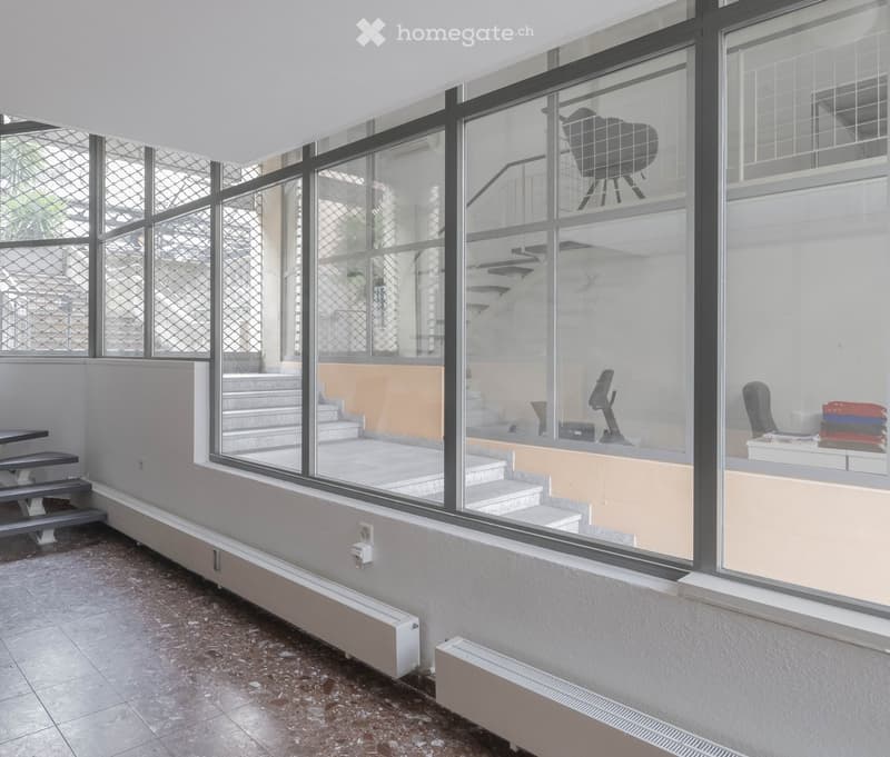 Empty room with tiled floor, large windows with metal grilles, radiator, and a small staircase leading to another area.
