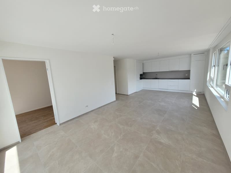 Empty living space with tiled floor, white walls, and a doorway leading to a kitchen area with white cabinets and countertops.