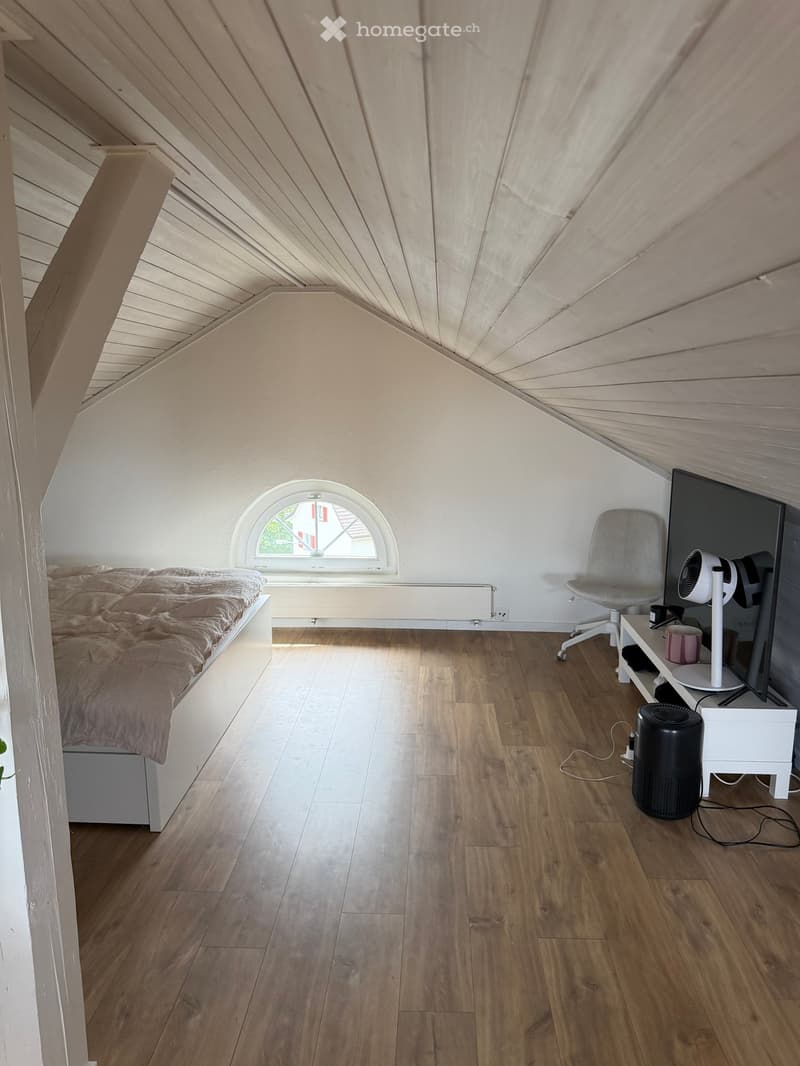 Cozy attic bedroom with wooden floors, slanted wooden ceiling, arched window, bed with beige bedding, grey chair, fan, and small table with screen.