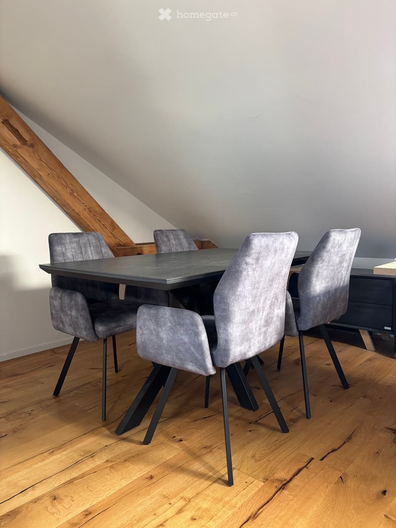 Dining area with a black dining table and four grey upholstered chairs on a wooden floor.