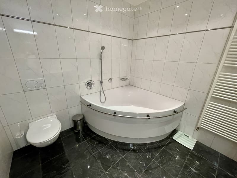 Modern bathroom with a white bathtub, toilet, and a chrome faucet. The walls are tiled in white with a dark marble floor.