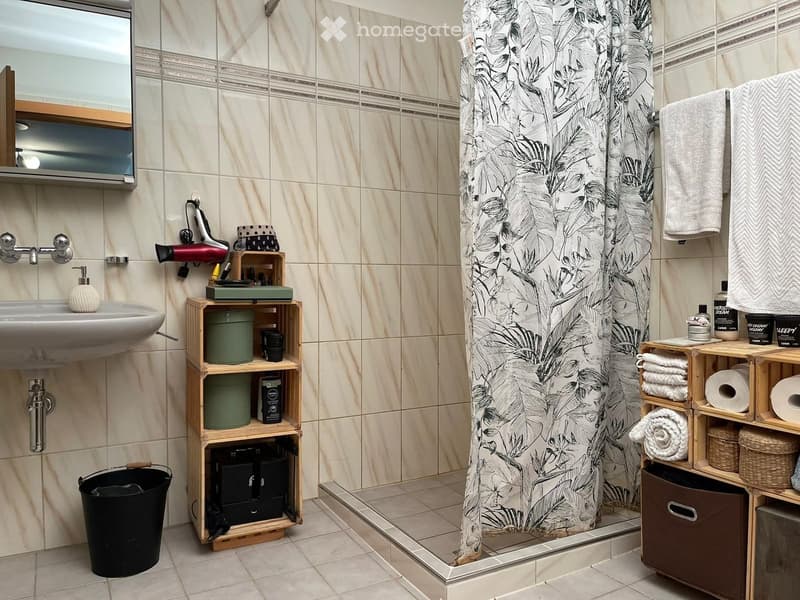 Bathroom with beige tiled walls, a sink with a mirror, a shower with a leaf-patterned curtain, and wooden shelves holding towels, toiletries, and other items.