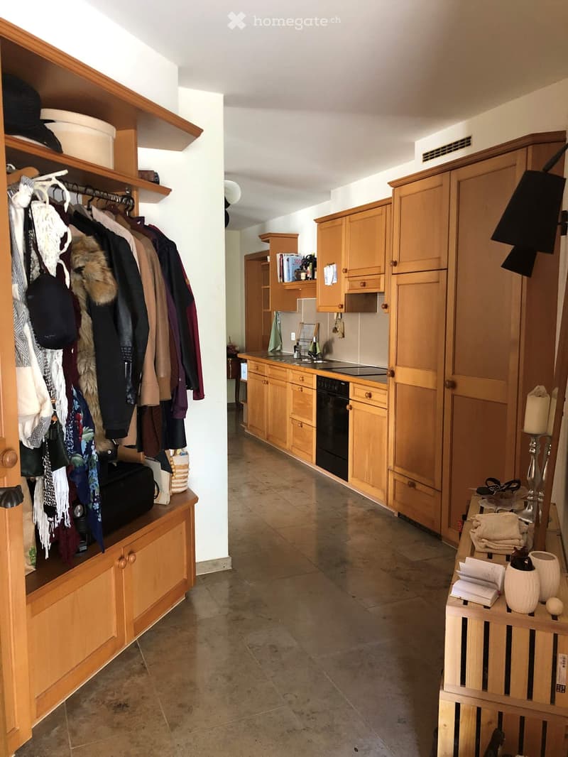 A well-lit hallway with wooden built-in storage units, a small kitchen area with wooden cabinets, and a tiled floor.
