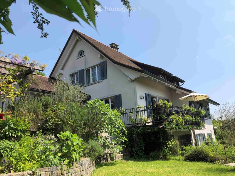 Two-story house with a pitched roof, multiple windows with shutters, a balcony, and surrounding greenery.