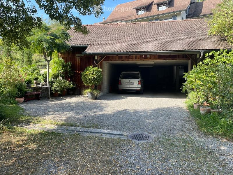 Two-story house with a large attached garage, gravel driveway, and surrounding greenery including trees and shrubs.