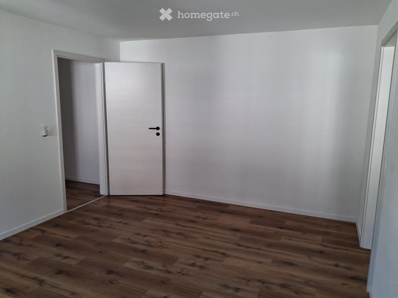 Empty room with wooden flooring and white walls, featuring a closed door and an electrical outlet visible on the wall.