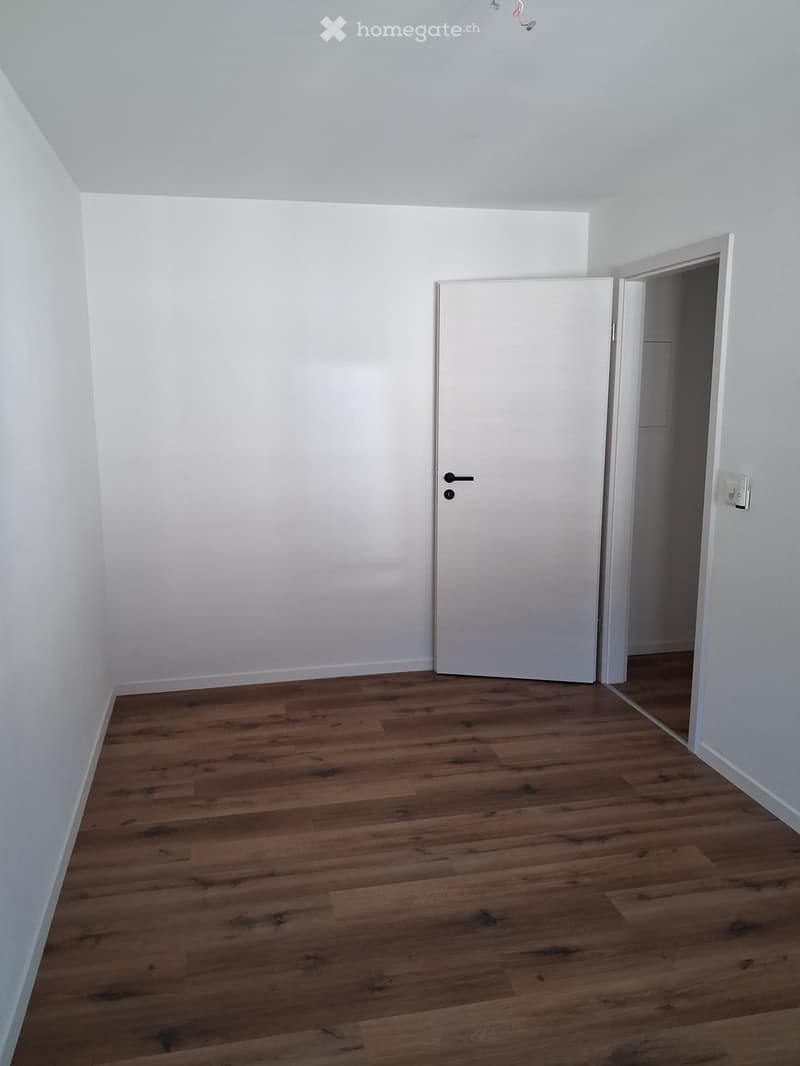 Empty room with wooden flooring and white walls, featuring a closed white door and an open doorway leading to another room.