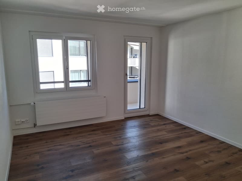 Empty room with wooden flooring, white walls, large windows, and a sliding glass door leading to a balcony.