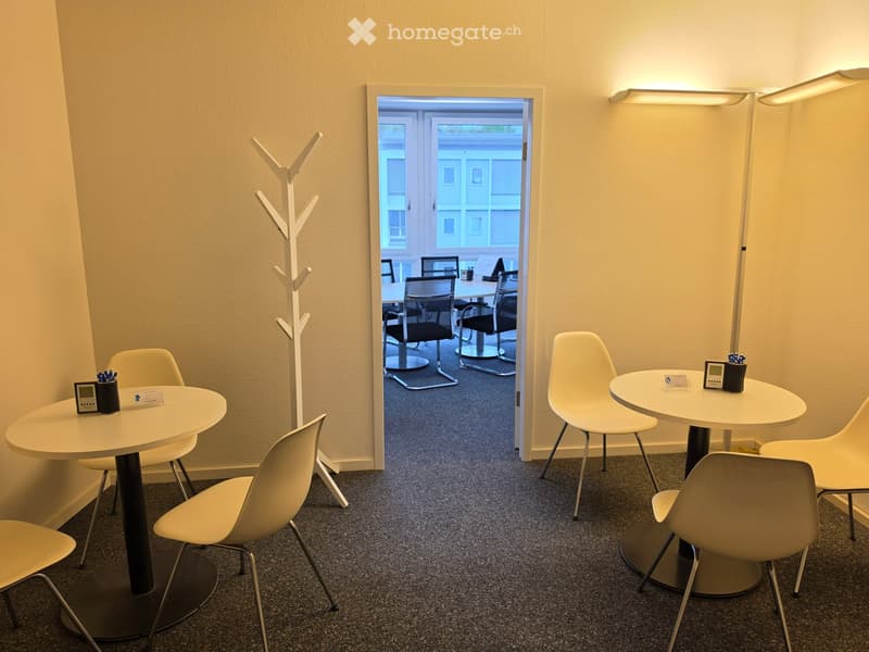 Modern, minimalist room with white walls, carpeted floor, and a coat rack. Two round tables with chairs are arranged, and a floor lamp is positioned near the doorway leading to another room.