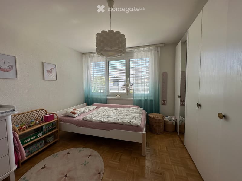 Cozy children's bedroom with a single bed, nightstand, round rug, and wicker baskets. Features large windows with curtains and decorative picture frames on the wall.