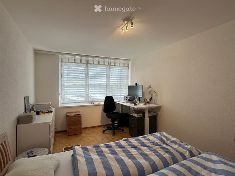 Cozy bedroom with a bed, desk, chair, and computer. Features a large window with blinds, a wicker basket, and a small cabinet.