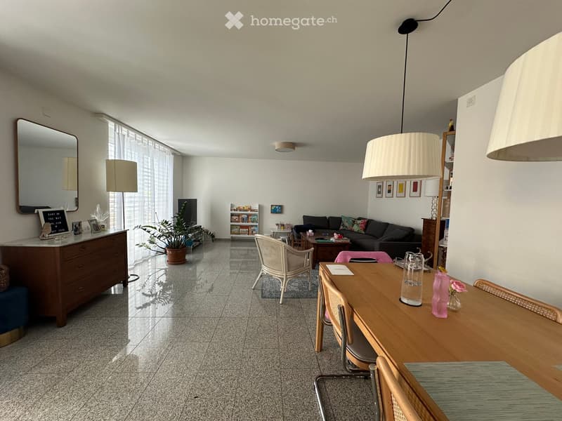 Bright living and dining area with a sofa, coffee table, dining table, chairs, and a large mirror. The space features modern decor, including lamps, a plant, and artwork on the walls.