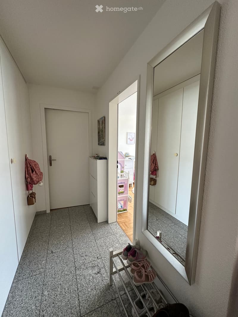 A narrow hallway with tiled flooring, white walls, and a slanted ceiling. Features include a small shoe rack with children's shoes, a mirror cabinet, a small storage unit, and a partially visible room with a toy playhouse.
