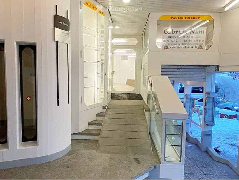 A clean, modern hallway with white walls, glass display cases, and a staircase leading upwards. Signage for 'Paulis Toyshop' and 'Gabriele Nani' is visible, along with a view of a parking area through a glass wall.