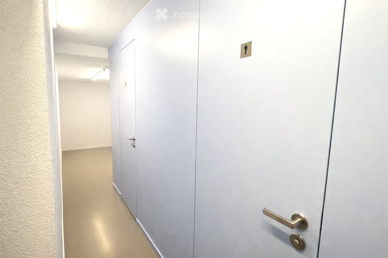A clean, well-lit hallway with white walls and a light-colored floor. Features include a closed door on the left and a restroom sign on the right wall.